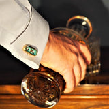 A hand pouring a drink from a decanter, where the subject is wearing sculptural bronze cufflinks